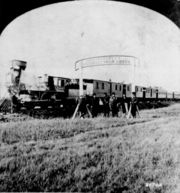 Directors of the Union Pacific Railroad gather on the 100th meridian, which later became Cozad, Nebraska, approximately 250 miles (400km) west of Omaha, Nebraska Territory, in October 1866. The train in the background awaits the party of Eastern capitalists, newspapermen, and other prominent figures invited by the railroad executives.